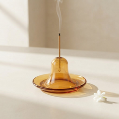 An amber glass bell-shaped incense holder with a single burning incense stick inserted, set on a light beige surface with soft shadows and a small white flower prop nearby.
