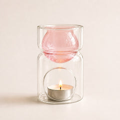 A clear glass candle holder with a pink glass top insert, shown with a small lit tealight candle inside on a warm off-white background.