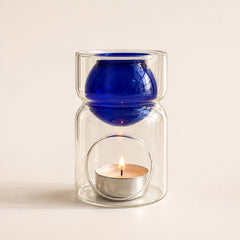 A clear glass candle holder with a stacked design, featuring a deep cobalt blue glass bowl on top and a silver tea light candle lit inside a clear glass jar below, set against a warm off-white background.
