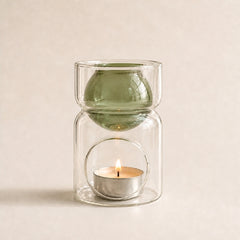 A double-walled glass candle holder with a green-tinted upper chamber and a clear lower chamber holding a lit tealight candle, set against a warm beige background.