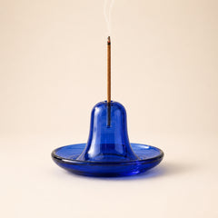 A blue glass incense holder with a bell-shaped center and a flat saucer base, shown with a lit incense stick standing upright inside. The holder is photographed on a plain warm beige background with a small wisp of smoke rising.