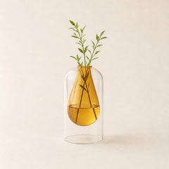 An amber glass dropper-style vase with a clear dome-shaped outer cover, holding water and green stems, centered on a warm off-white background.