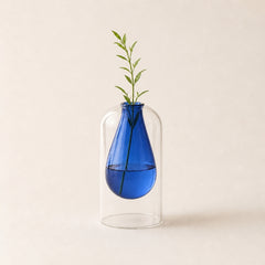 A small cobalt blue glass vase suspended inside a larger clear glass dome, holding a single green sprig, on a warm off-white background.
